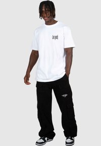 Vit bomull t-shirt med "lost youth" grafik på vänster bröst, ihop med svarta cargobyxor med sidofickor och vingad logotyp.
