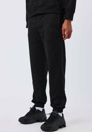 Pantalon de jogging noir en tissu doux et texturé avec une taille élastique, poches latérales et un discret logo. Associé à des baskets noires.