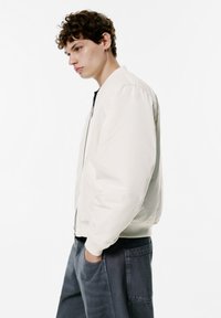 Light beige bomber jacket with elastic cuffs, front pockets, and a zip closure. Worn over dark gray pants, featuring a plain design.
