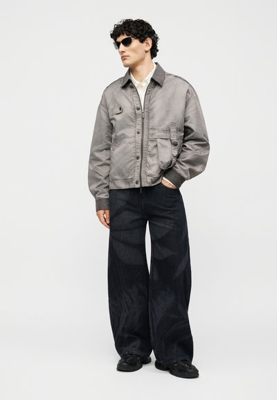 Άνδρας που φορά γκρι μπουφάν, λευκό πουκάμισο, μαύρα τζιν με φαρδιά μπατζάκια, μαύρα αθλητικά παπούτσια και μαύρα γυαλιά ηλίου, στέκεται με το ένα χέρι στην τσέπη.