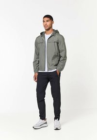 Kevyt, oliivinvihreä hupparillinen takki, jossa on vetoketjullinen edusta ja joustavat hihansuut, yhdistettynä mustiin kapeisiin housuihin ja harmaisiin lenkkareihin.