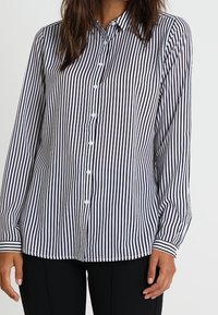 Chemise à rayures noires et blanches avec un col, des manches longues et des boutons blancs, présentant une coupe droite et des manches à poignets.
