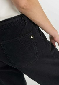 Pantalones de pana negra con un bolsillo trasero que presenta un pequeño detalle en dorado. La tela tiene una textura acanalada y un corte recto.