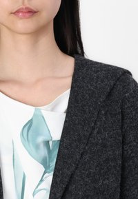 Dark gray, textured wool coat with a shawl collar, paired with a white top featuring a light teal abstract pattern.