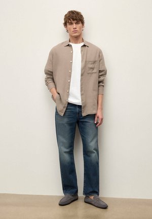 Jeune homme debout contre un mur uni, portant une chemise beige à boutons par-dessus un t-shirt blanc, un jean bleu et des mocassins gris.