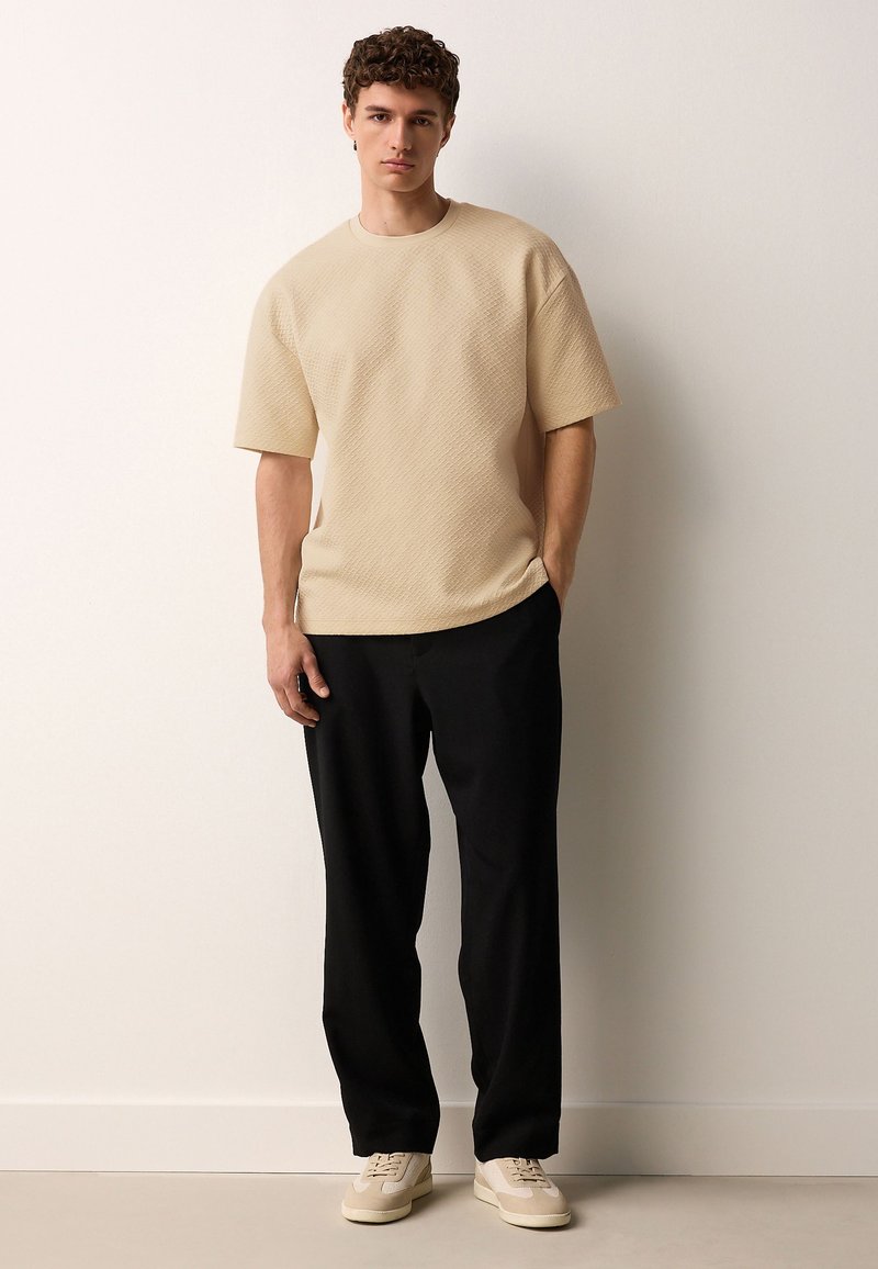 Beige short-sleeve textured shirt paired with black pants and light-colored sneakers. Simple design with relaxed fit and round neckline.