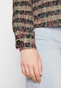 Tweed jacket with a multicolored pattern in green, red, and black. Features a buttoned cuff with a dark round button. Casual fit.