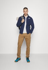 Mørkeblå fleece zip-up jakke, hvid T-shirt, beige cargo-bukser og blå mønstrede træningssko. Rygsæk med beige stropper.