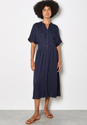 Kiharatukkainen nainen, jolla on päällään tummansininen napillinen midi-mekko ja beigevät tasapohjaiset sandaalit, seisomassa valkoista seinää vasten.