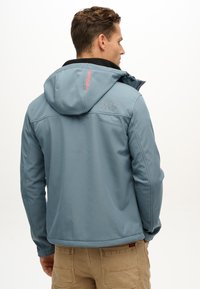 Veste softshell bleue avec capuche, doublure intérieure noire, logo "SUPERDRY" en gris et accents rouges, au design ajusté.