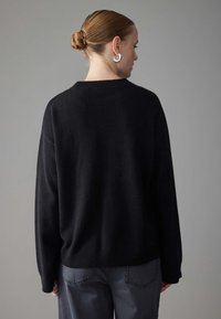 Black knit sweater with a relaxed fit, ribbed neckline and hem, paired with gray trousers. Sweater has dropped shoulders and long sleeves.
