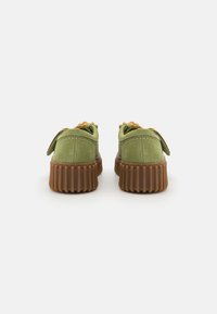 Zapatillas de ante verdes con una suela de goma marrón texturizada, presentando cordones naranjas y un diseño redondeado con una pestaña en el talón.