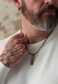 Collier en cuir noir avec un pendentif métallique rectangulaire. Le pendentif présente une petite accent en diamant. Le porteur a des tatouages sur sa main.
