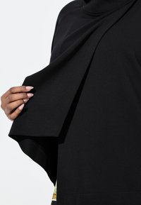 Czarny strój o gładkiej, miękkiej fakturze, z warstwowym wzorem. Widać dłoń trzymającą fałdę, ukazującą układ i materiał.