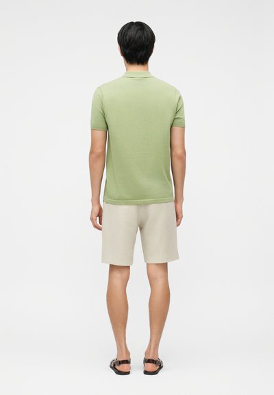 Homme de dos portant un polo à manches courtes vert clair, un short beige et des sandales noires sur un fond blanc uni.