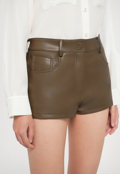 Barna műbőr rövidnadrág magas derekú, elülső zsebekkel és látható varrási részletekkel; fehér gombos blúzzal párosítva.
