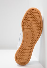 Vedere de jos a unui sneaker Reebok cu o talpă din cauciuc maro, prezentând un model texturat pentru aderență și branding în relief; materialul superior alb este parțial vizibil.