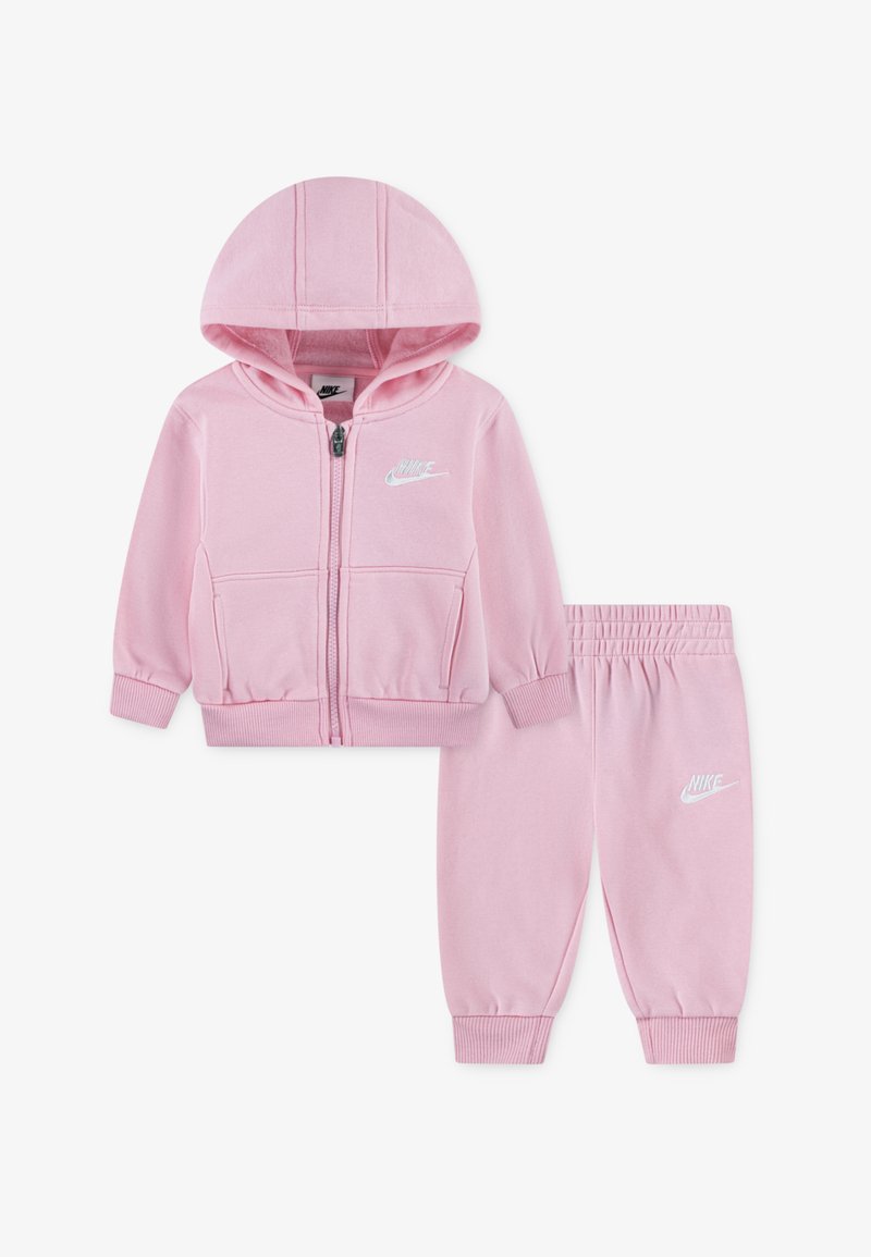 Ροζ σετ φούτερ και φόρμες της Nike. Το φούτερ διαθέτει φερμουάρ και μπροστινές τσέπες· και τα δύο κομμάτια έχουν λευκά λογότυπα Nike και ριμπ μανσέτες.