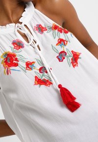 Weißes ärmelloses Blusenshirt mit gerüschtem Ausschnitt, verziert mit buntem Blumenstickerei in Rot, Blau und Grün, sowie einem roten Quastenschmuck.