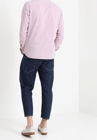 Hellrosa Cord-Hemd mit abgerundetem Saum, kombiniert mit dunkelblauen, eng zulaufenden Jeans und grauen Wildlederschuhen mit weißen Sohlen.