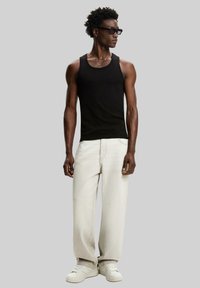 Débardeur noir, jean large beige clair, et baskets blanches. Modèle avec des cheveux courts et texturés et des lunettes de soleil noires. Éléments de design simples.