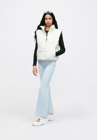 Λευκό αμάνικο μπουφάν με ψηλό γιακά, κλείσιμο με φερμουάρ και πλαϊνές τσέπες, φορεμένο πάνω από μακρυμάνικη μαύρη μπλούζα και συνδυασμένο με ανοιχτόχρωμο μπλε παντελόνι με καμπάνα.