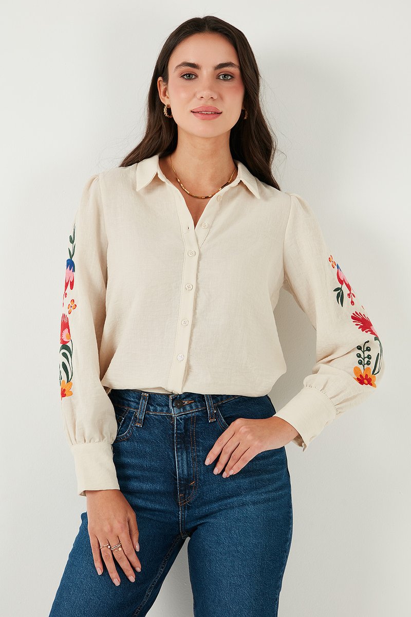 Blusa crema de manga larga con bordado floral en las mangas, cierre de botones en la parte delantera y corte relajado, combinada con unos jeans oscuros.