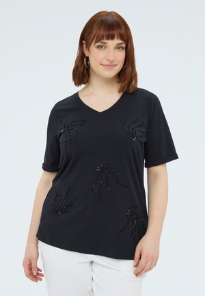 Femme aux cheveux roux portant des boucles d’oreilles créoles, un haut noir à manches courtes orné de perles et un pantalon blanc, debout devant un fond clair uni.