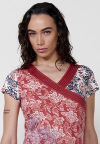 Rotes Blumenshirt mit V-Ausschnitt und kurzen Ärmeln, aus gemustertem Stoff mit blauen und weißen Akzenten sowie strukturierten Rippdetails am Ausschnitt.