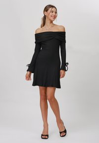 Abito nero a spalla scoperta con texture a costine, maniche lunghe, orlo svasato e dettagli con fiocco nei polsini, realizzato in un tessuto elastico.