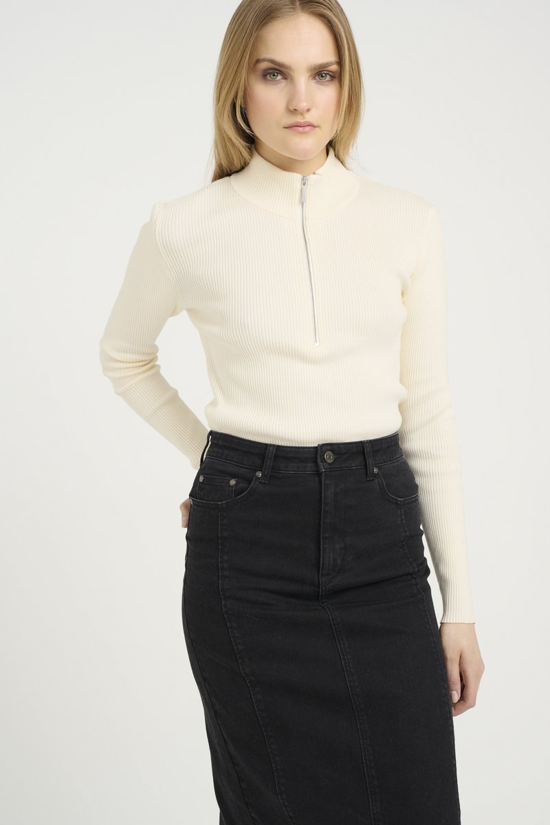 Maglione a coste di colore crema con collo alto e chiusura frontale a mezza zip. Abbinato a una gonna in denim nera a vita alta, con tasche.