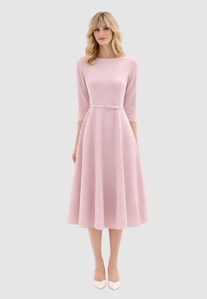 Donna con capelli biondi mossi che indossa un vestito midi rosa chiaro con cintura, maniche a tre quarti e décolleté bianche con punta appuntita, in piedi davanti a uno sfondo grigio.