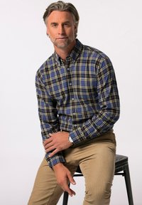 Camisa de cuadros en tonos azules y amarillos, de manga larga, fabricada en un tejido suave. Pantalones de pana beige. Modelo sentado en un taburete negro.