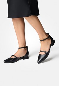 Ballerines en cuir noir avec une bride à la cheville, bout pointu, petit talon et découpes uniques sur les côtés. Texture lisse.