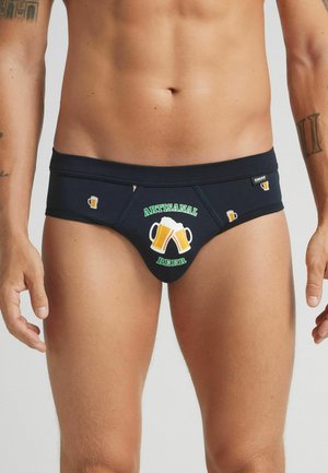 Dunkelblaue Herrenunterhosen mit bedruckten Bierkrügen und dem Text "ARTISANAL BEER" auf der Vorderseite, getragen von einem tätowierten männlichen Model vor einem schlichten Hintergrund.