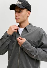 Hombre con gorra negra y camisa gris con cremallera abrochando la cremallera con ambas manos contra un fondo claro y uniforme.