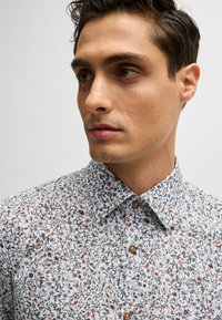 Chemise à imprimé floral sur fond blanc, ornée de petites fleurs roses et bleues, avec un col classique et des boutons marron. Texture lisse.