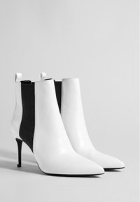 Botines blancos de tobillo con punta afilada, cuerpo de cuero y paneles laterales de tela negra. Presentan un delgado tacón de aguja y tiradores para facilitar su puesta.