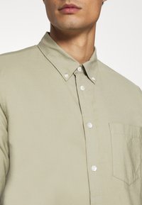 Homme portant une chemise boutonnée vert olive clair avec une poche et un col boutonné, visible du menton à la poitrine.