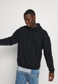 Μαύρη φούτερ με μπροστινή τσέπη, ρυθμιζόμενο καρουλάκι και αντίθετες λεπτομέρειες στα μανίκια, σε συνδυασμό με ανοιχτό μπλε τζιν παντελόνι.