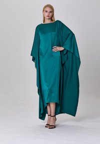 Robe kaftan en satin turquoise avec un col bateau et des manches légèrement évasées. Elle présente une texture lisse et arrive au niveau du mollet, associée à des talons noirs.