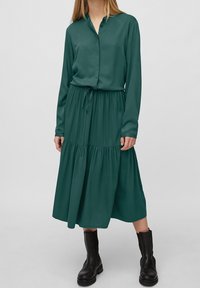 Pitkähihaista vihreää mekkoa, jossa on napitettava kaulus, rypistetty vyötärö ja kerroksellinen hame. Yhdistetty mustien nilkkabootsien kanssa. Suora pinta.