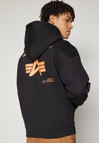Black hoodie featuring an orange logo design on the back, with graphic text detailing and soft, textured fabric. Hooded with long sleeves.