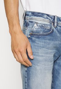 Jeans de mezclilla en azul claro con un acabado desgastado. Cuenta con un bolsillo para monedas, remaches de metal y costuras visibles en la cintura y los laterales.