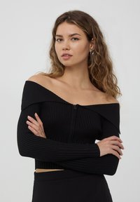 Cardigan cropped nero a coste con design off-shoulder, maniche lunghe e zip frontale. Presenta un materiale testurizzato e una forma aderente.