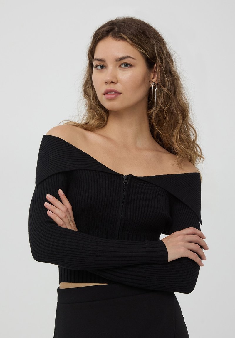 Cardigan cropped nero a coste con design off-shoulder, maniche lunghe e zip frontale. Presenta un materiale testurizzato e una forma aderente.