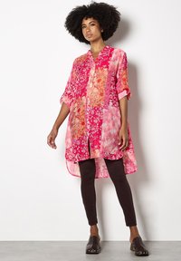Roze tuniek met patroon, voorzien van een knoopsluiting aan de voorkant en een high-low zoom, met bloem- en abstracte ontwerpen. Gecombineerd met donkere leggings en klompen.