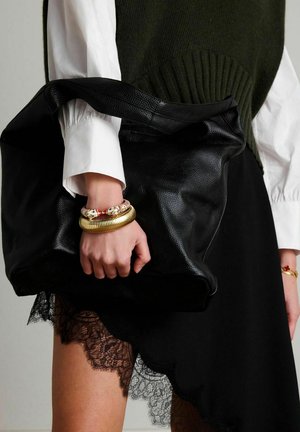 Zwarte leren handtas met een gestructureerde afwerking, vastgehouden door een hand met meerdere armbanden. De outfit bestaat uit een wit overhemd en een groen gebreid vest.