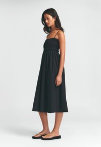 Vestido negro de largo medio con un cuerpo ajustado fruncido y tirantes delgados, combinado con sandalias deslizantes color tostado. Tejido suave y diseño sencillo.