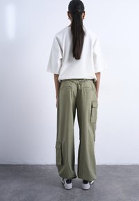 Γυναίκα με μακριά σκούρα μαλλιά δεμένα πίσω, φορώντας άνετο λευκό πουκάμισο, χακί παντελόνι cargo και λευκά αθλητικά παπούτσια, όρθια σε τσιμεντένιο δάπεδο.